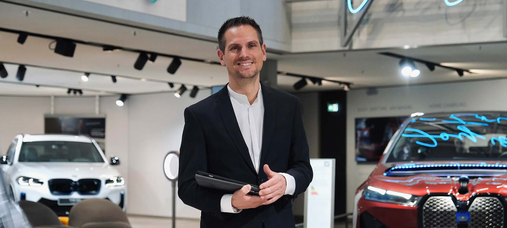 BMW Welt Fahrzeugberatung Lächelnder Mann im Anzug hält Tablet für die Fahrzeugberatung in der BMW Welt.
